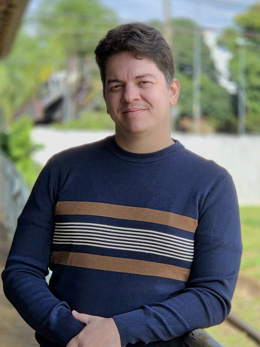 Dr. Hugo Hoffmann, de suéter azul-marinho listrado, sorrindo e apoiado em um corrimão ao ar livre.